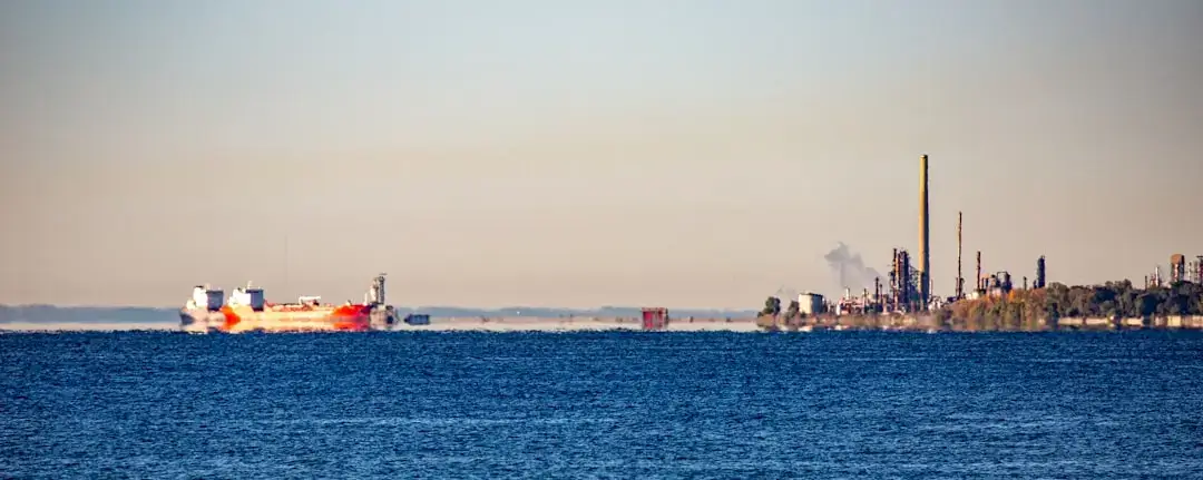 An industrial crude oil refinery featuring metal pipes, distillation towers, and steam silhouetted against a dramatic sunset sky.