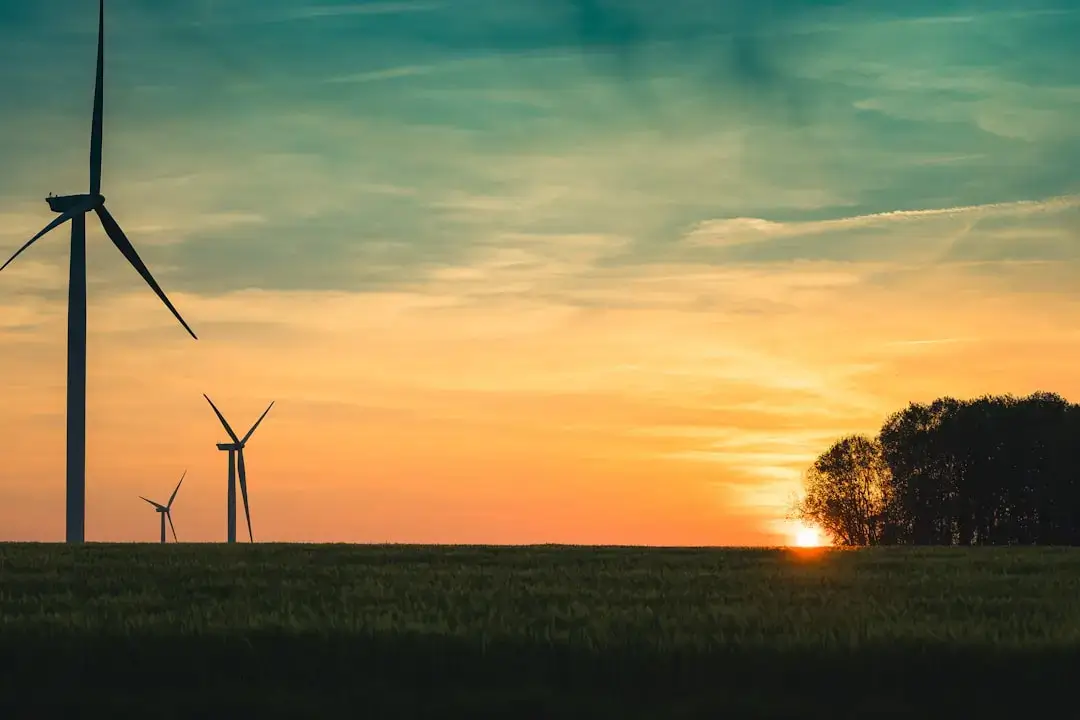 A line of modern white wind turbines standing in a lush green rolling field under a clear blue sky.